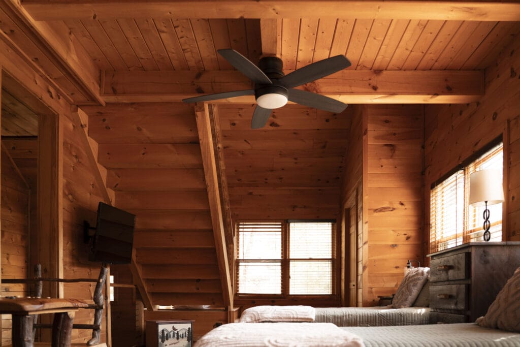 One of the rooms of the Lincoln Logs Cabin, with a small desk, television, drawer, 2 beds, a ceiling fan, and plenty of sunlight